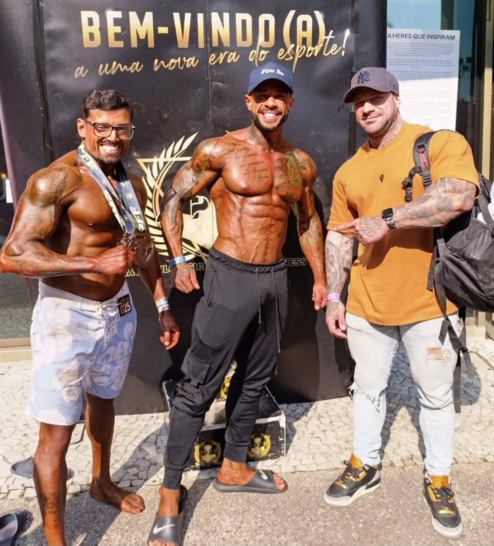 Joel de camiseta amarela ao lado de dois atletas, um deles com medalhas