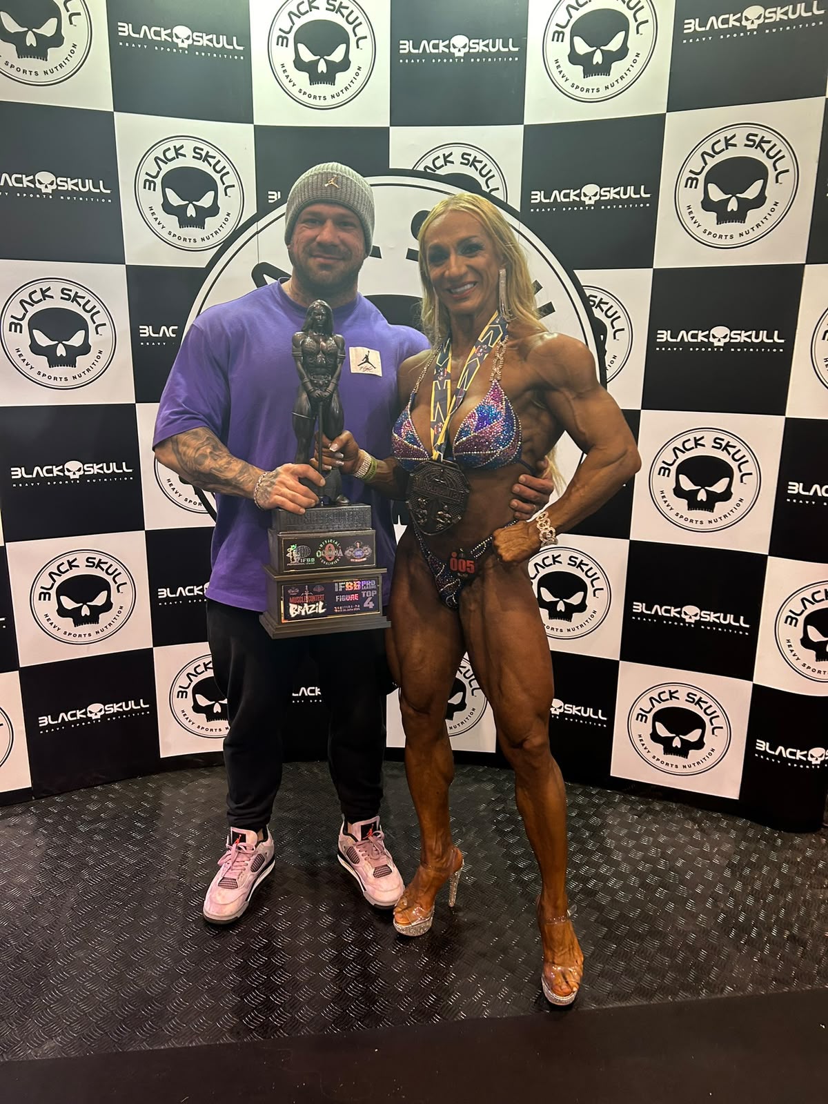 Joel de camiseta roxa ao lado de uma atleta com medalhas e troféu